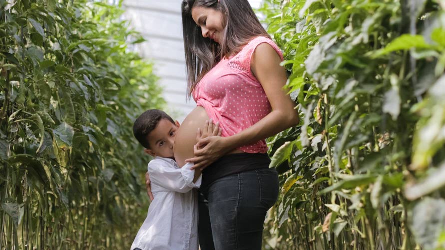 A pregnant woman stands in a field of plants with a child