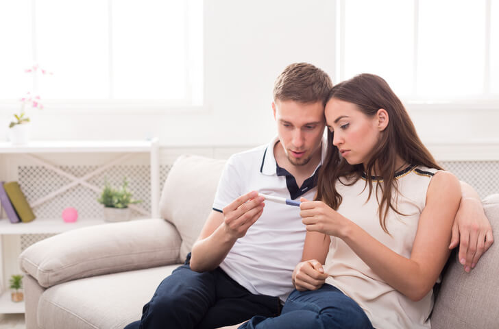a couple sitting on a beige couch looking at a pregnancy test