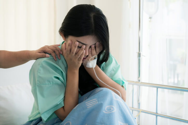  A woman sits on a hospital bed covering her face with her hands, her partner’s hand rests on her shoulder.