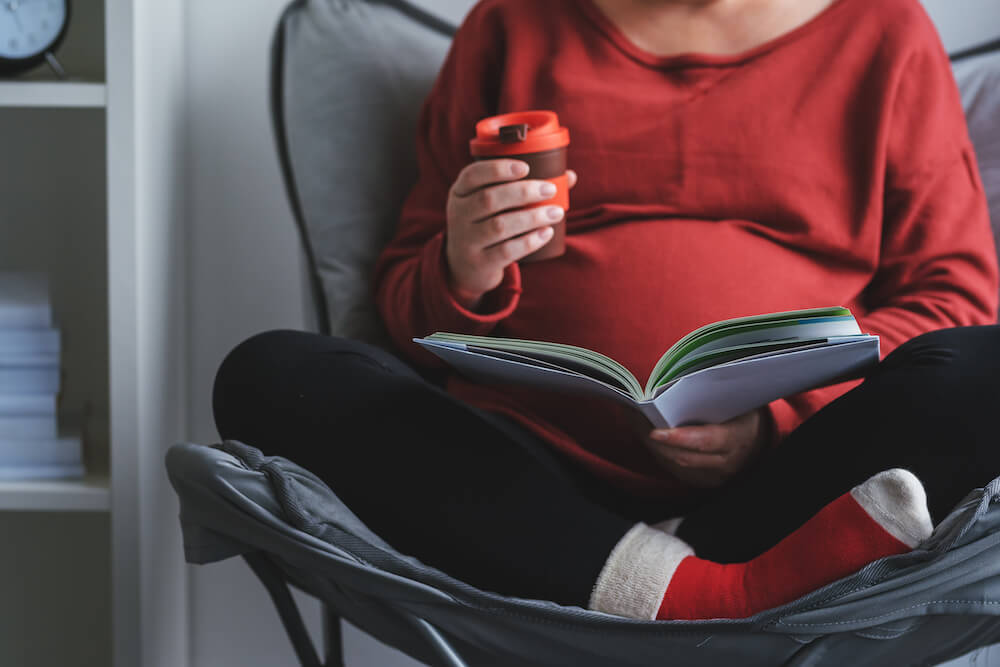 a pregnant woman reading a book and drinking from a coffee cup