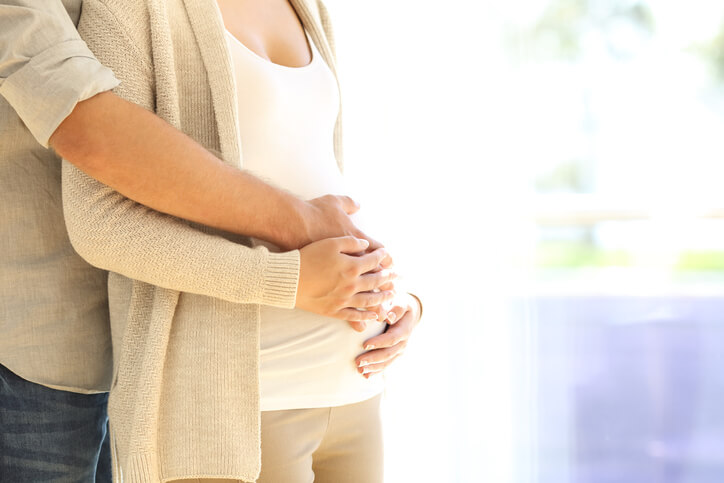 A pregnant couple stands together with their hands on the woman’s belly.