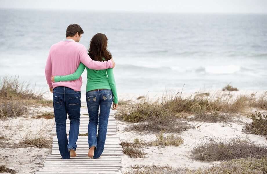 A couple walks arm-in-arm along a beach.